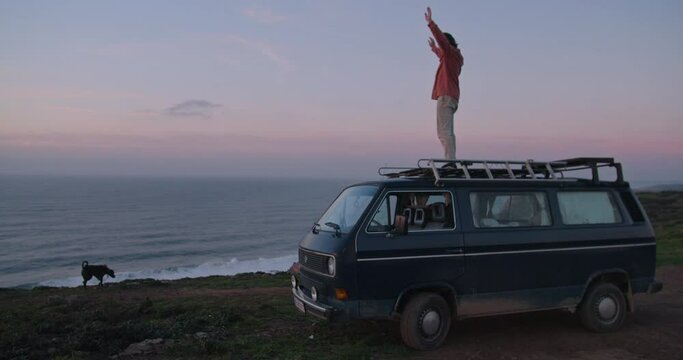 Young man watching and celebrating the sunset on the roof of his vintage van during golden hour along the cliffs and coastline of the Western Algarve, Portugal