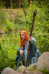 A young woman in a cloak, with a staff and a green dress in the form of a druid. Forest witch with...