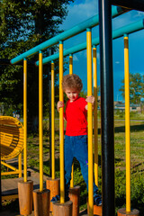 A curly-haired little boy in a red T-shirt walks on the playground. The child develops in the process of sports