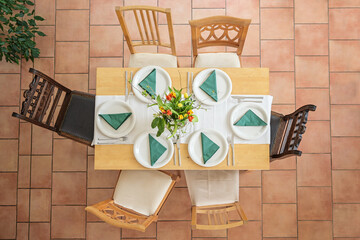 Wooden table with place setting for six with white plates, cutlery, green napkins and a flower bouquet, different vintage chairs on a tiled terracotta floor, high angle view from above