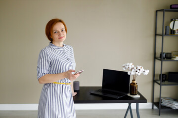 Fototapeta na wymiar red haired woman in striped dress against background of beige wall, holds smartphone in her hand and looks at the camera. Distant work. Online training. Home office