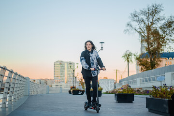A young man with long hair of an informal appearance rides along the embankment on an electric scooter. Guy in autumn in the city