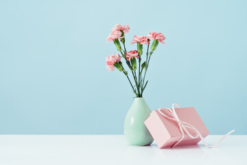 Pink gift box with tender pink carnation flowers in vase. Blue background. Gift or holiday concept. Mothers Day, birthday wedding or St Valentines day with copy space. Minimal