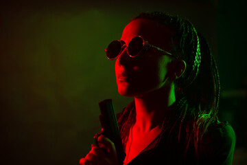 A young woman with pigtails in the dark in a club in round dark glasses holds a gun in her hands....