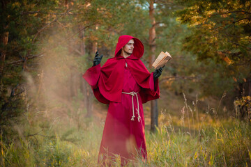 A monk in a red cassock with a book in his hands is praying in the forest. Male magician conjures in nature.