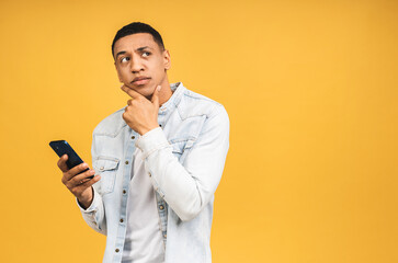 Serious thinking african american young man holding phone isolated on yellow studio background with copy space aside, african guy using mobile applications, texting.