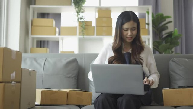 Female entrepreneur working at home office with smartphone with laptop, tablet, deck (SME business). Business concept of Asian woman checking customer orders at home, delivering goods to customers.