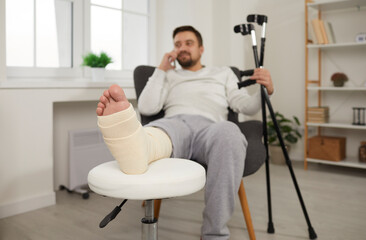 Unhealthy young man is sitting at home in chair with broken leg in cast and talking on the phone....