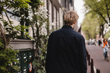 Portrait of blonde man wear black shirt walk in city street. Young man look from back, rear view....