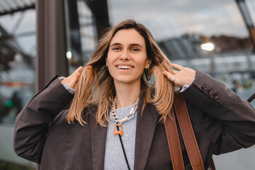 Attractive happy woman wear gray jacket, blue sweater, brown bag and necklace standing on bus station, fix her hair look flirty. Blonde woman waiting bus look at camera.