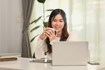Obraz premium Charming asian female entrepreneur watching online webinar, communicating on internet via laptop computer
