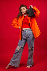 A young girl in a bright orange shirt and jeans on a red background holds a tape recorder from the...