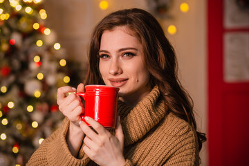 A young woman in a beige sweater sits in a Christmas decoration against the backdrop of a Christmas tree and lights. The girl is holding a red mug in her hands.