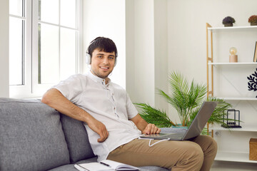 Cheerful male student freelancer at home watching online training or webinar on laptop. Young man in headphones listens to information while sitting on sofa with notebook and pen.