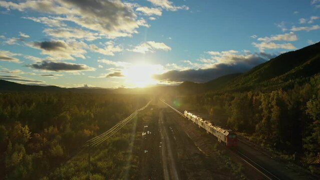 The train leaves into the sunset in this mesmerizing drone video. As the train slowly chugs along the tracks, the sun sets behind it, casting a warm, golden glow over the surrounding landscape. The sk