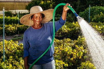 Black woman wearing a straw hat and using a garden hose to water the plants