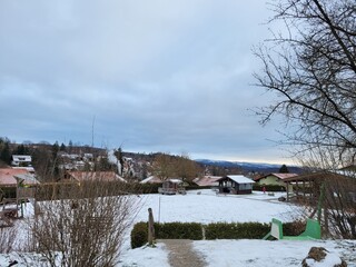 Houses and trees in a hilly landscape covered with snow with a cloudy sky and mountains in the background
