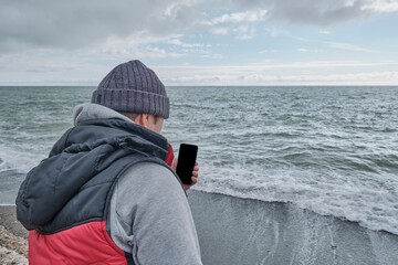 Senior man using cellphone with black empty screen for mockup, outdoors near sea.