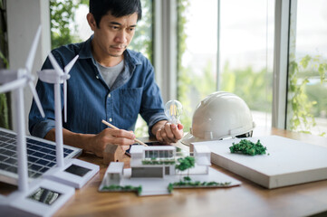 An engineer sits holding a light bulb and a tablet looking at a model of a house. with solar panels...