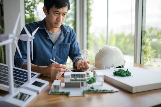 An engineer sits holding a light bulb and a tablet looking at a model of a house. with solar panels and a model house on the table. To design use of renewable energy with wind and solar energy.