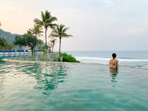 A young woman leaning at the edge of an infinity pool and looking at the sea. 