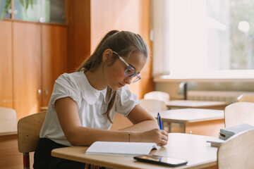 teenage girl in glasses reading in copybook at desk in classroom