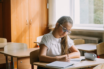 teenage girl in glasses reading in copybook at desk in classroom