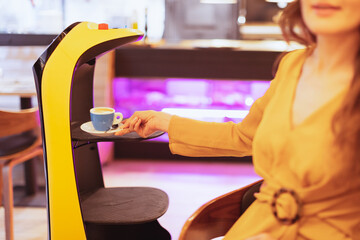 Picture of robot serving coffee to a customer in a restaurant. Young woman holding a cup of espresso coffee served by a robot.