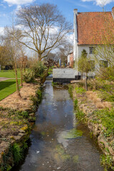 Thornerbeek stream and old Kraekermolen watermill in background, reflection on water surface, house with white walls and bare trees against blue sky, sunny day in Thorn, Midden-Limburg, Netherlands
