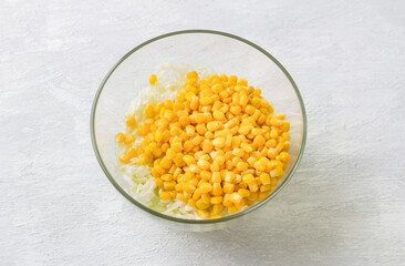 Glass bowl with chopped white cabbage and sweet corn on a light gray background, top view. Stage of cooking healthy vegan salad