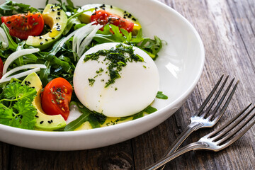 Burrata cheese with leafy greens, avocado and cherry tomatoes on wooden table
