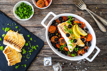 Cod in Italian style on tomatoes and black olives with baked potatoes in baking dish on wooden table

