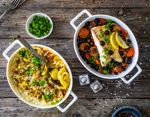 Cod in Italian style on tomatoes and black olives with baked potatoes in baking dish on wooden...