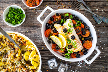 Cod in Italian style on tomatoes and black olives with baked potatoes in baking dish on wooden table

