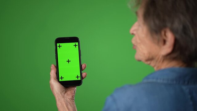 Over the shoulder view of elderly woman talking on smart phone with green screen tracking points isolated on green screen background with mock up copy space. Hold mobile phone with blank empty screen