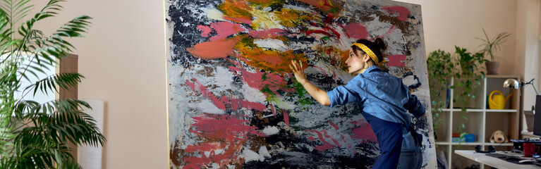 Full length shot of inspired female painter working on a large modern abstract oil painting, applying paint on canvas with fingers, at home studio workshop