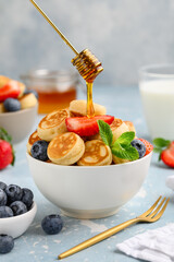 Mini pancakes in a bowl  with strawberries and blueberries. Flowing honey on a stick. Trendy tiny pancake. Healthy breakfast, snack, dessert. Selective focus