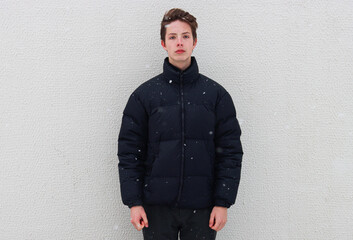 A belted portrait of a teenage boy in a black jacket against a wall in wintertime. A boy against a background of falling snowflakes.