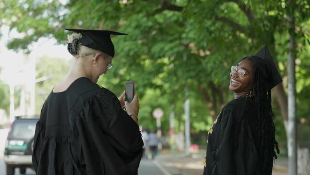 Caucasian White Female University Student takes pictures of her Black Colleague with smartphone for social media after receiving degrees. Celebrate academic success after Graduation.