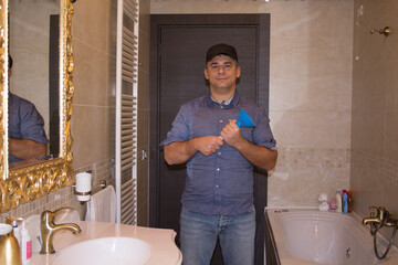 Image of a smiling plumber in the home bathroom holding a plunger in his hand while unclogging a...