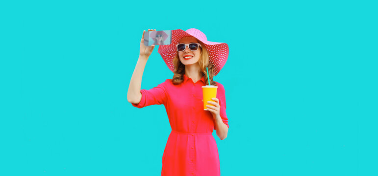 Summer portrait of beautiful happy smiling young woman taking selfie with smartphone and cup of juice wearing straw hat, pink dress on blue background