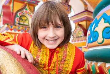a happy boy in a Russian national bright red-orange costume near the Russian national multi-colored Terem building.
