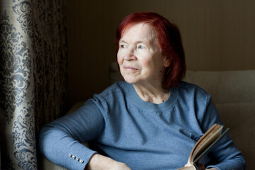 Portrait of elderly woman with open book in hands. Senior woman spending time reading paper book, at home, sitting on sofa. old woman with red hair, thoughtful, looking out window. Life in old age.