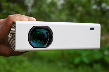 A White Projector with green Shiny Lens in Hands at Outdoor, It's a Chinese LED projector front...