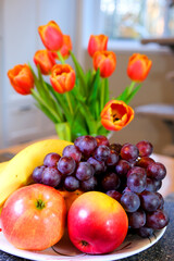 room on table apples grapes bananas red pink tulips with yellow edging cat sits in the background white kitchen cabinets against the window beautiful home furnishings
