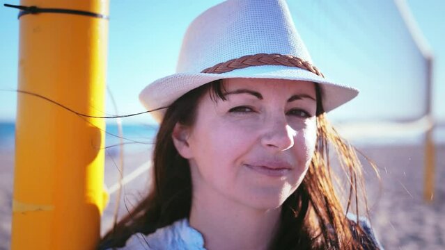 Slow-motion shot of a happy woman in a stylish hat standing near a volleyball net, enjoying the beautiful surroundings and the moment