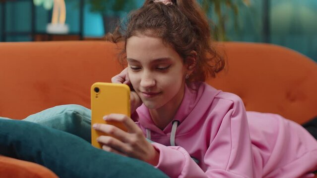Preteen school girl texting share messages content on smartphone social media applications online, watching relax movie. Happy child kid uses mobile phone at home room apartment lying on orange couch