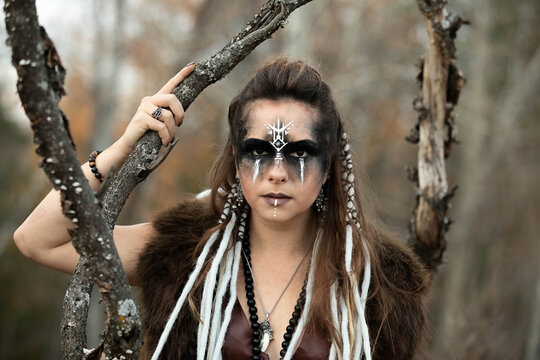 Autumn portrait of a female viking wearing face paint, braids, leather and fur. She is standing in the forest. 