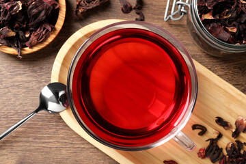Cup of fresh hibiscus tea, dry flower leaves and spoon on wooden table, flat lay