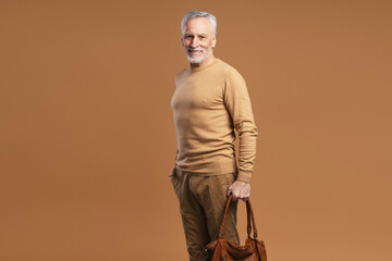 Smiling elderly man standing isolated on brown background holding leather bag. Elderly male